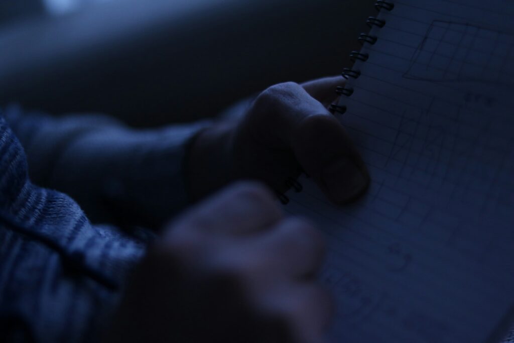 A close-up photo of a person taking notes in a spiral notebook under dim blue lighting, possibly during nighttime study or work.