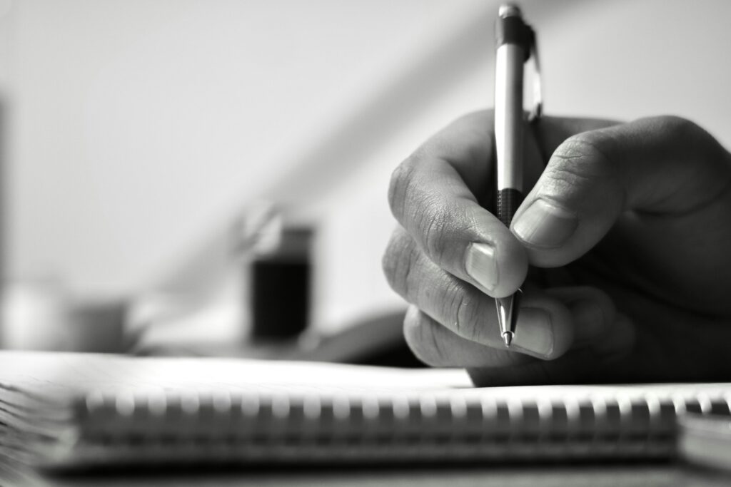 Close-up of a hand holding a pen, writing on a spiral notebook, in black and white.
